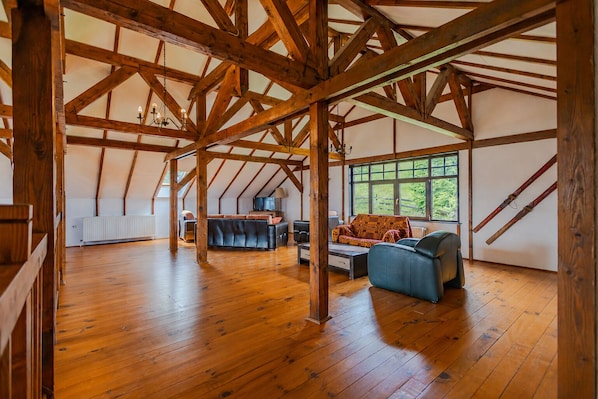 A spacious attic room with exposed wooden beams, a large window with a view of greenery, and a cozy seating area with a sofa and chairs.