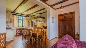 A dining area with a wooden table and chairs, a kitchen in the background, and a wooden door.