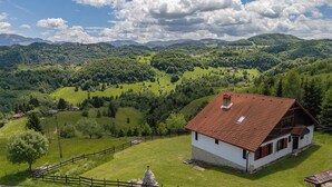 A rural house with a red-tiled roof, surrounded by lush green fields and mountains in the background.