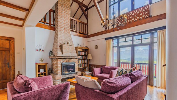A spacious living room with a fireplace, wooden beams, and a balcony with a view.