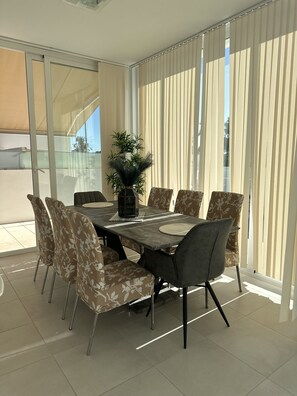 Comedor con una mesa rectangular, seis sillas, una planta en maceta y ventanas grandes con persianas verticales.