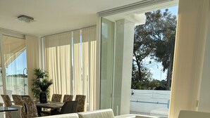 Una sala de estar moderna con un sofá seccional color beige, una zona de comedor con una mesa de vidrio y ventanas grandes con cortinas.