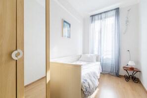 Een slaapkamer met een bed, nachtkastje, lamp, raam met gordijnen en een aan de wand bevestigd schilderij.