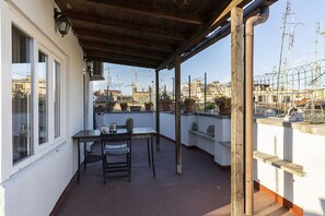 Een overdekt balkon buiten met een tafel, stoelen, potplanten en uitzicht op de stadsgezicht.