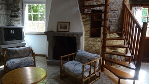 Une pièce chaleureuse avec une cheminée en pierre, un escalier en bois, une télévision et deux fauteuils en osier.