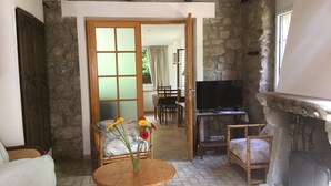Un salon chaleureux avec un mur en pierre, une cheminée, un téléviseur et un coin repas visible à travers une porte.