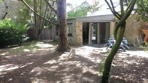 Un jardin avec un mur en pierre, un arbre et un espace patio aménagé avec des chaises.