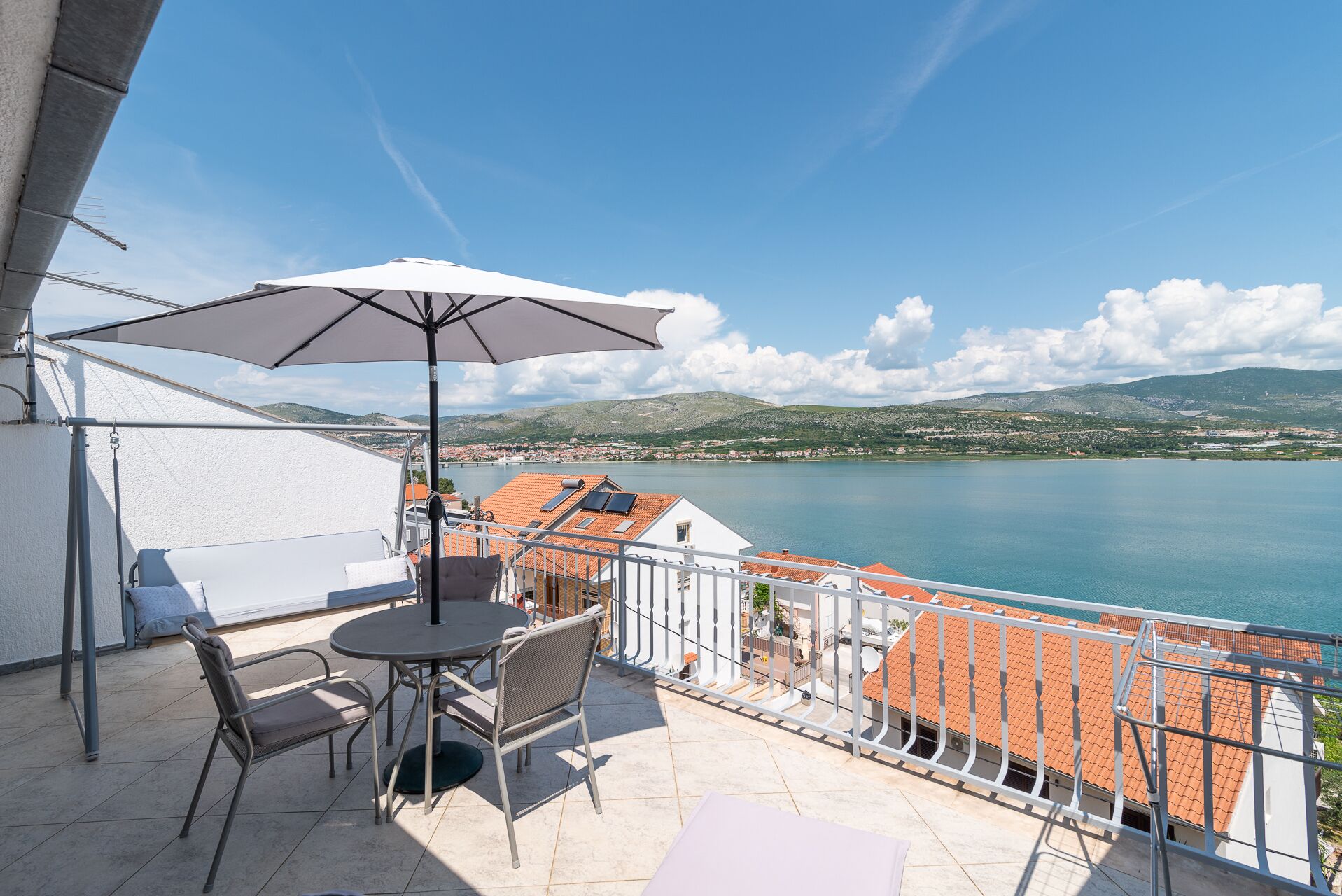 A balcony with a table and chairs, an umbrella, and a view of a lake and distant hills.