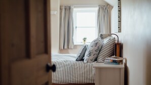 A bedroom with a bed, a bedside table with books, a lamp, and curtains. - Gorgeous flint cottage overlooking the marshes, walking distance to beach (Nr Cley next the Sea)