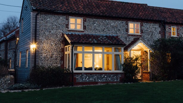 A stone house with a tiled roof, a porch with a glass door, and a well-lit interior. - Gorgeous flint cottage overlooking the marshes, walking distance to beach (Nr Cley next the Sea)