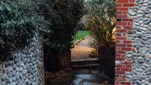 A stone pathway leading through a garden area with a brick wall on one side and a stone wall on the other. - Gorgeous flint cottage overlooking the marshes, walking distance to beach (Nr Cley next the Sea)