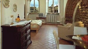 A room with a bed, a dresser, a chair, and a table with a vase. There are two windows with arched tops and a brick wall on the right. - King Suite - Michelangelo (Arezzo)