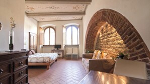 A spacious bedroom with a large bed, a wooden dresser, a sofa, and a stone wall. - King Suite - Michelangelo (Arezzo)