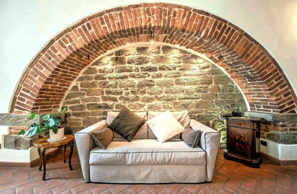 A cozy living room with a stone fireplace, a beige sofa, and a small wooden table with a plant. - King Suite - Michelangelo (Arezzo)