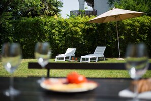 Une table avec une assiette de nourriture et deux fauteuils blancs, placés sous un grand parasol, le tout sur fond de végétation luxuriante et d’un bâtiment.