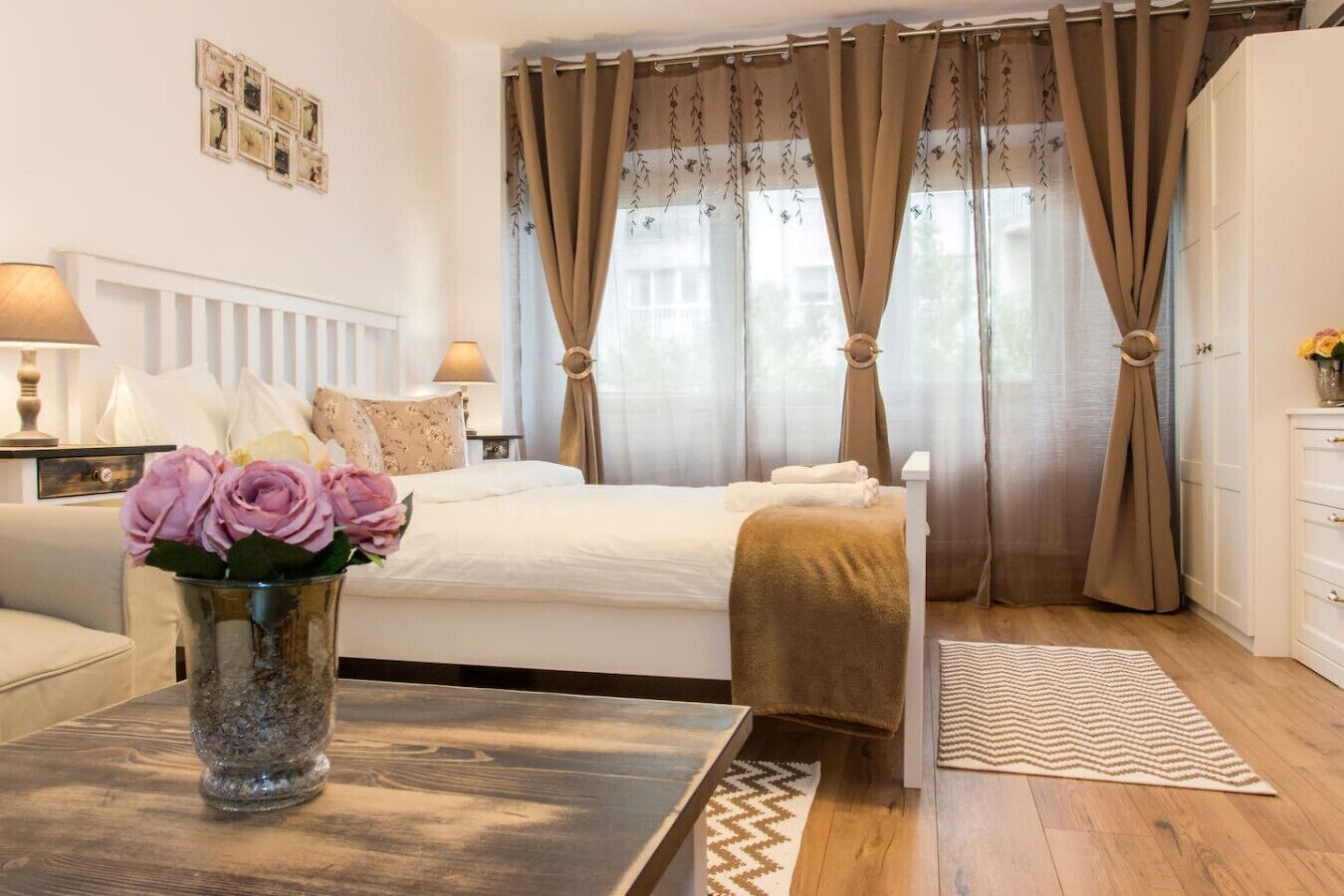Une chambre à coucher avec un lit, une table de chevet ornée d’un vase de fleurs, une commode et une fenêtre avec des rideaux.