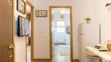 A hallway leading to a kitchen with a refrigerator, a dining table with a vase of flowers, and a wall-mounted TV.
