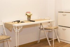 Una habitación pequeña con un escritorio blanco, dos sillas, un florero con flores y un armario blanco.