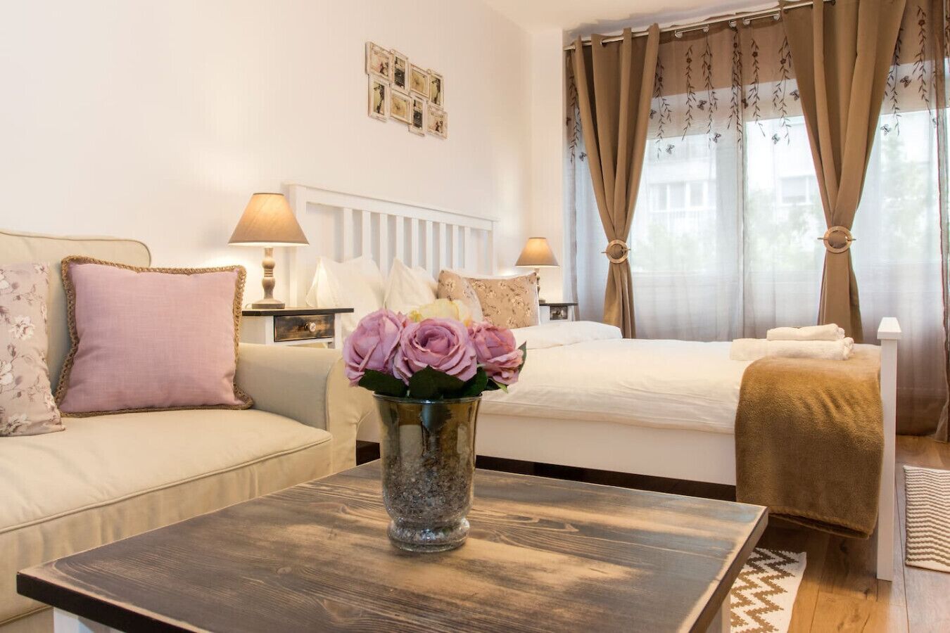 A hotel room with a bed, a sofa, a coffee table with a vase of flowers, and a window with curtains.