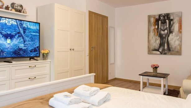 A hotel room with a bed, a television, a wardrobe, a painting, and a small table with flowers. - Motru 75 by MRG Apartments | Chic & Spacious Retreat in Central Area (București)