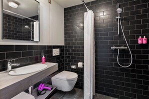 A modern bathroom with a black tiled wall, a white sink, a toilet, and a shower with a curtain. - Moxy Frankfurt Airport Kelsterbach (Kelsterbach)