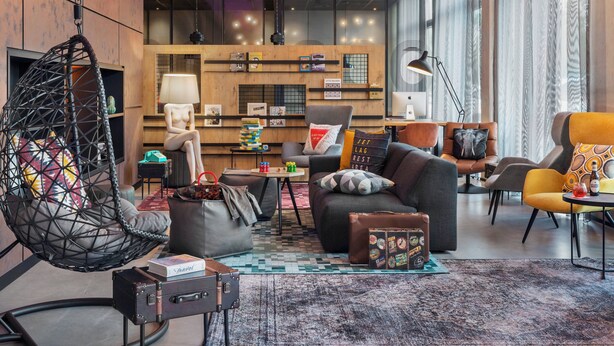 A modern lounge area with a hanging chair, sofas, a small table, and a suitcase. - Moxy Frankfurt Airport Kelsterbach (Kelsterbach)