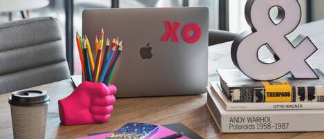 A laptop with colorful pencils, a coffee cup, a notebook, and a book titled "Andy Warhol Polaroids 1958-1987" on a wooden table.