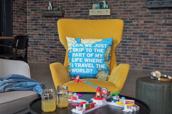 Un salon chaleureux avec un fauteuil jaune, une table basse décorée de jouets sur le thème du voyage et un coussin en forme de carte géographique.