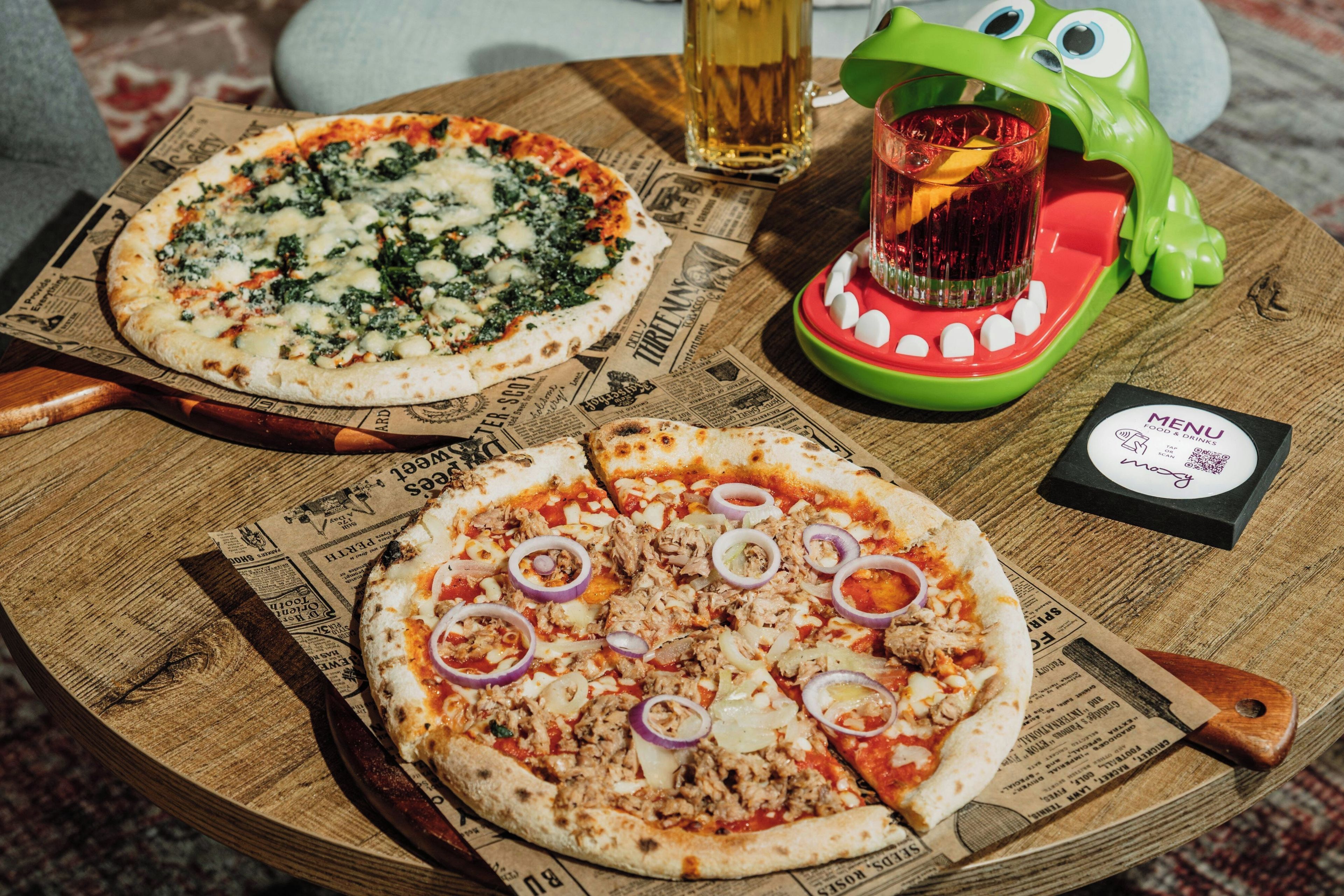 Zwei Pizzen auf einer Holzplatte, dazu eine Speisekarte und ein Getränk auf einem Tisch.