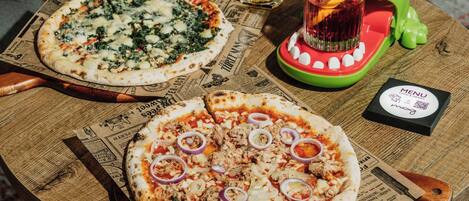 Two pizzas on a wooden board with a menu and a drink on a table.