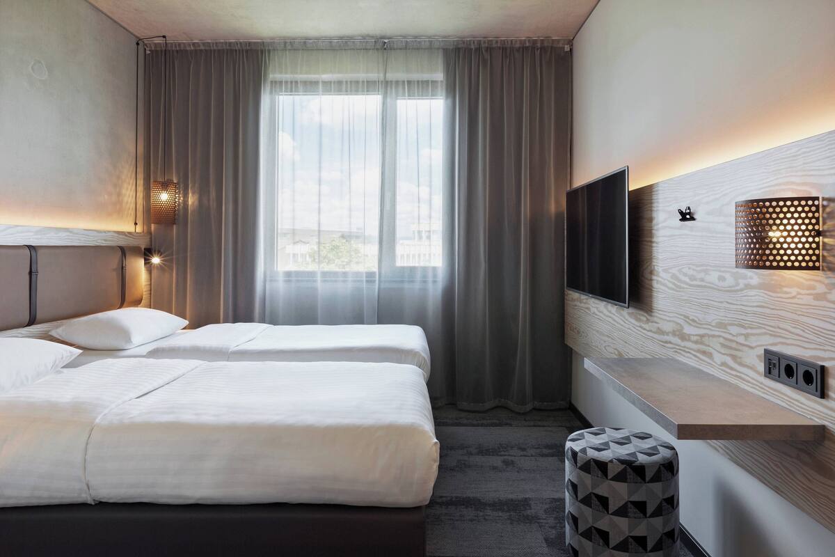 A hotel room with two beds, a TV, a wooden accent wall, and a window with curtains.