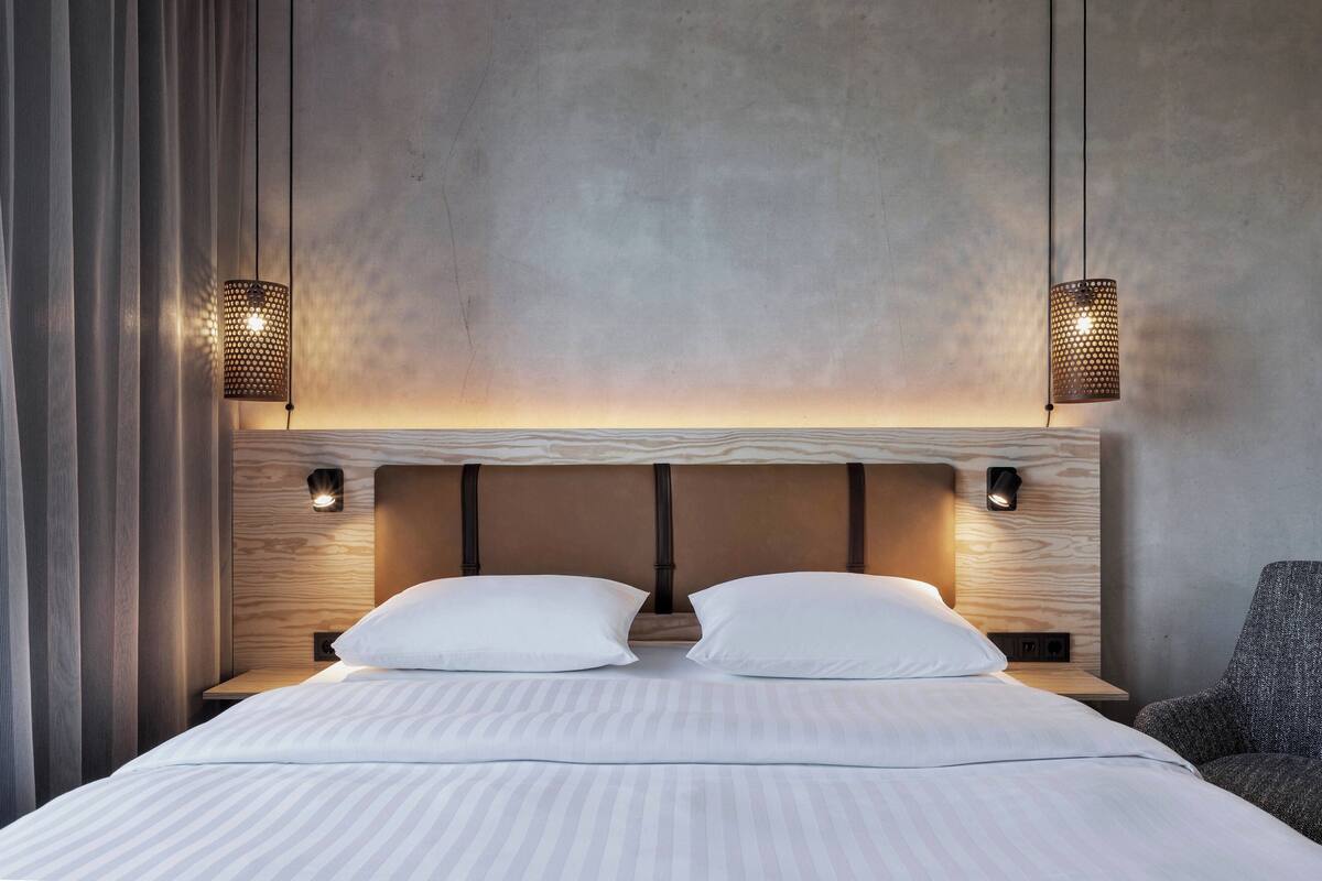A modern hotel room with a large bed, two wall-mounted lamps, and a view of the sky through the window.