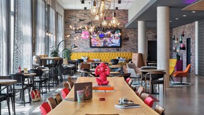 A modern dining area with a long wooden table, red chairs, and a large screen displaying a menu. - Moxy Frankfurt Airport Kelsterbach (Kelsterbach)