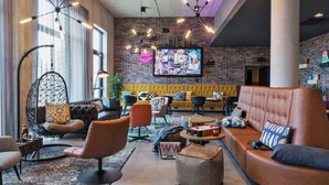A modern lounge area with a brick wall, comfortable seating, and a large screen displaying content. - Moxy Frankfurt Airport Kelsterbach (Kelsterbach)