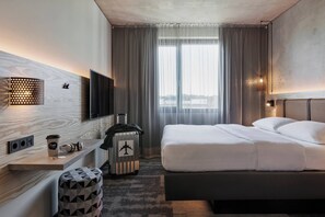 A hotel room with a bed, a desk, a chair, a suitcase, and a television. - Moxy Frankfurt Airport Kelsterbach (Kelsterbach)