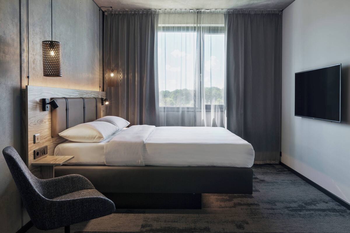 A modern hotel room with a bed, a chair, a TV, and a window with curtains.