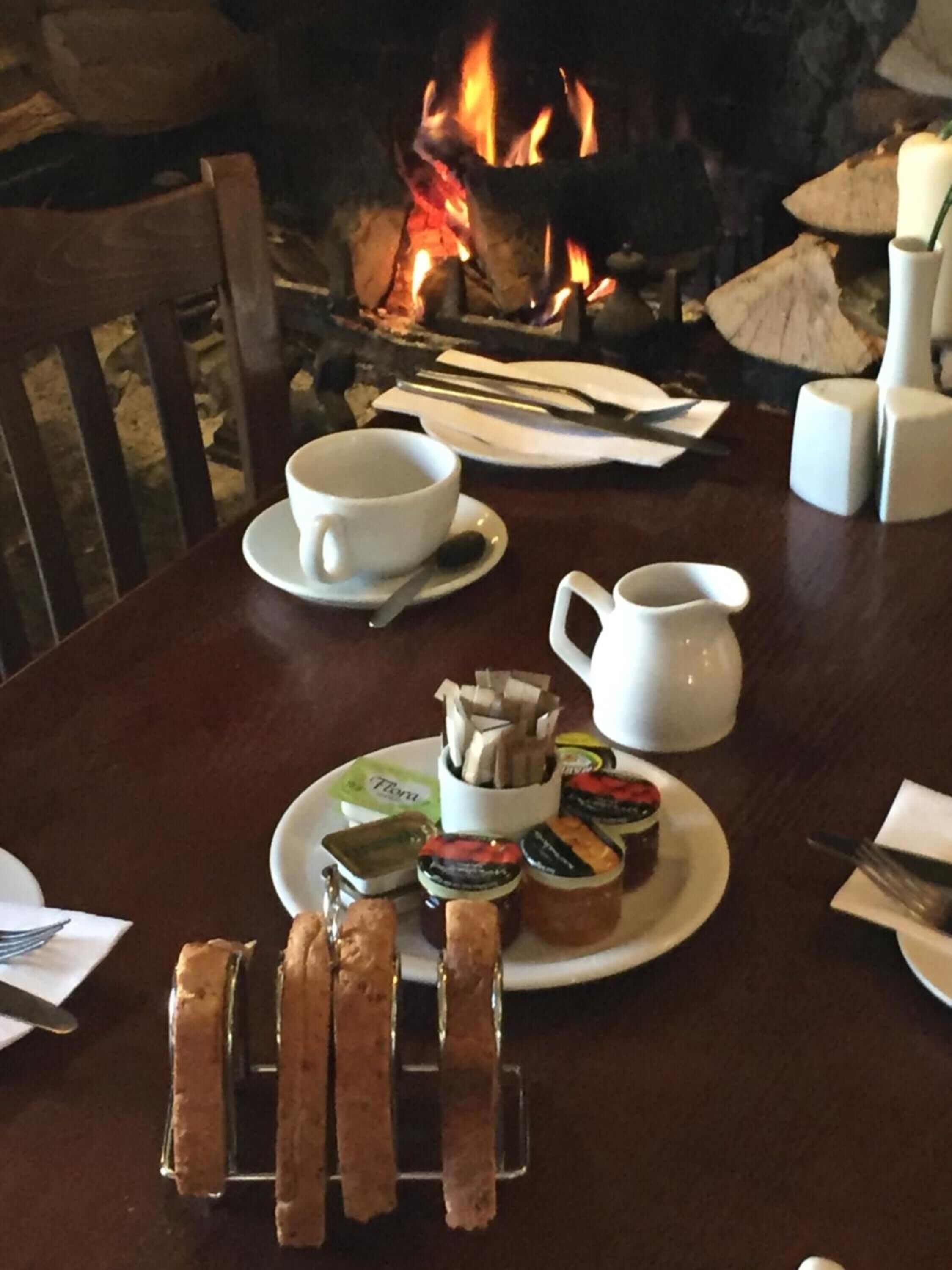 A dining table set for two with a plate of assorted jams, a white pitcher, and a cup of tea. A fire is visible in the background.