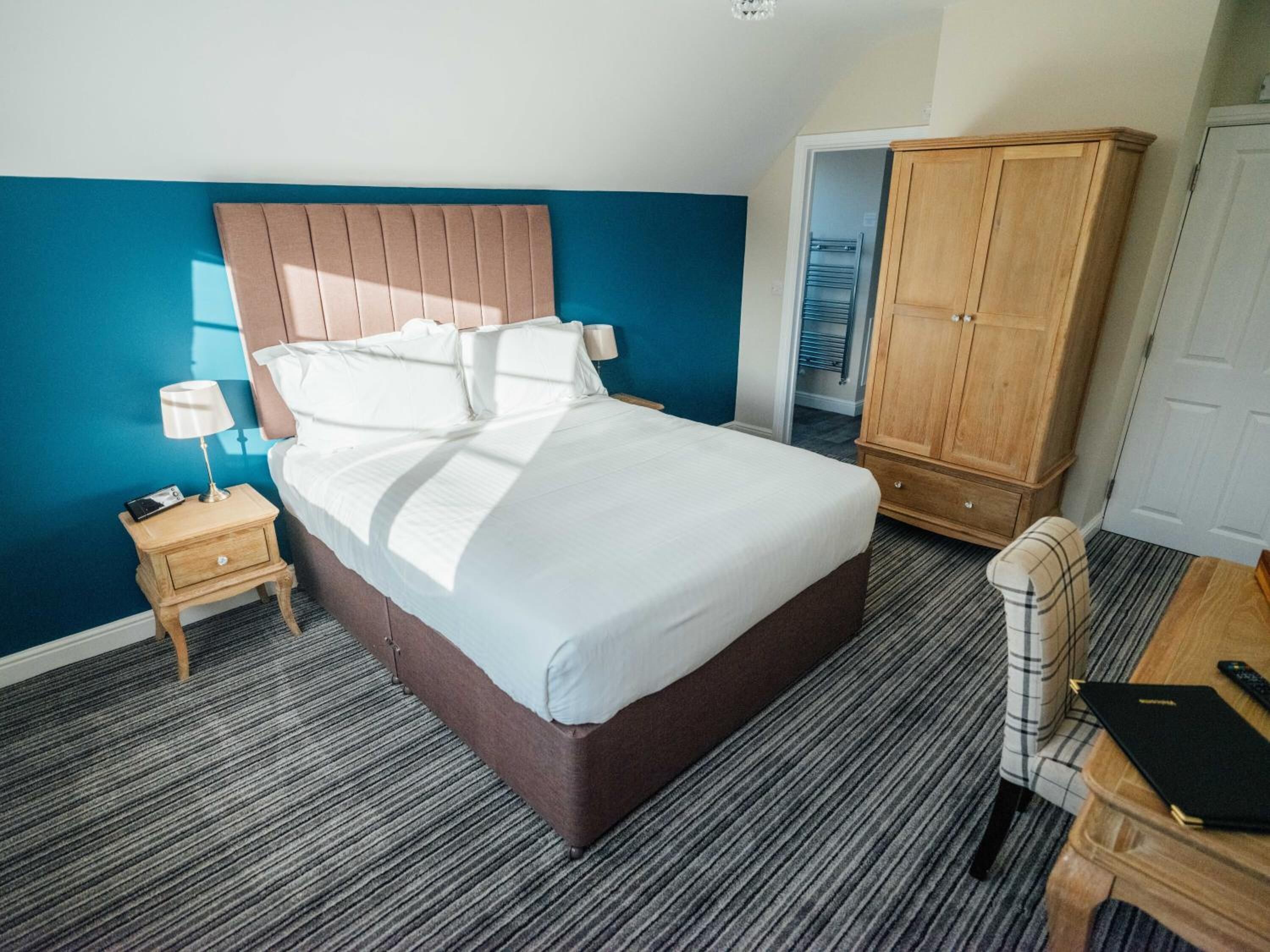 A hotel room with a large bed, a wooden wardrobe, a nightstand with a lamp, and a chair.