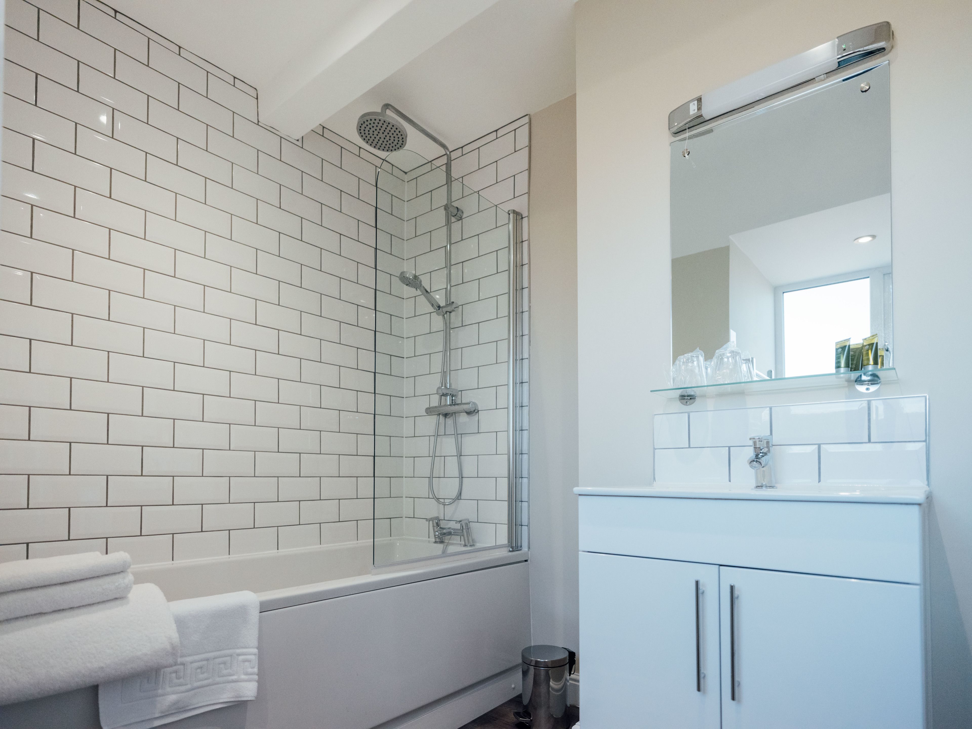 Un baño moderno con paredes de azulejos blancos, cabina de ducha con mampara de vidrio, bañera blanca y tocador con espejo y estante de vidrio.