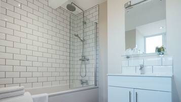 Un baño moderno con paredes de azulejos blancos, cabina de ducha con mampara de vidrio, bañera blanca y tocador con espejo y estante de vidrio.