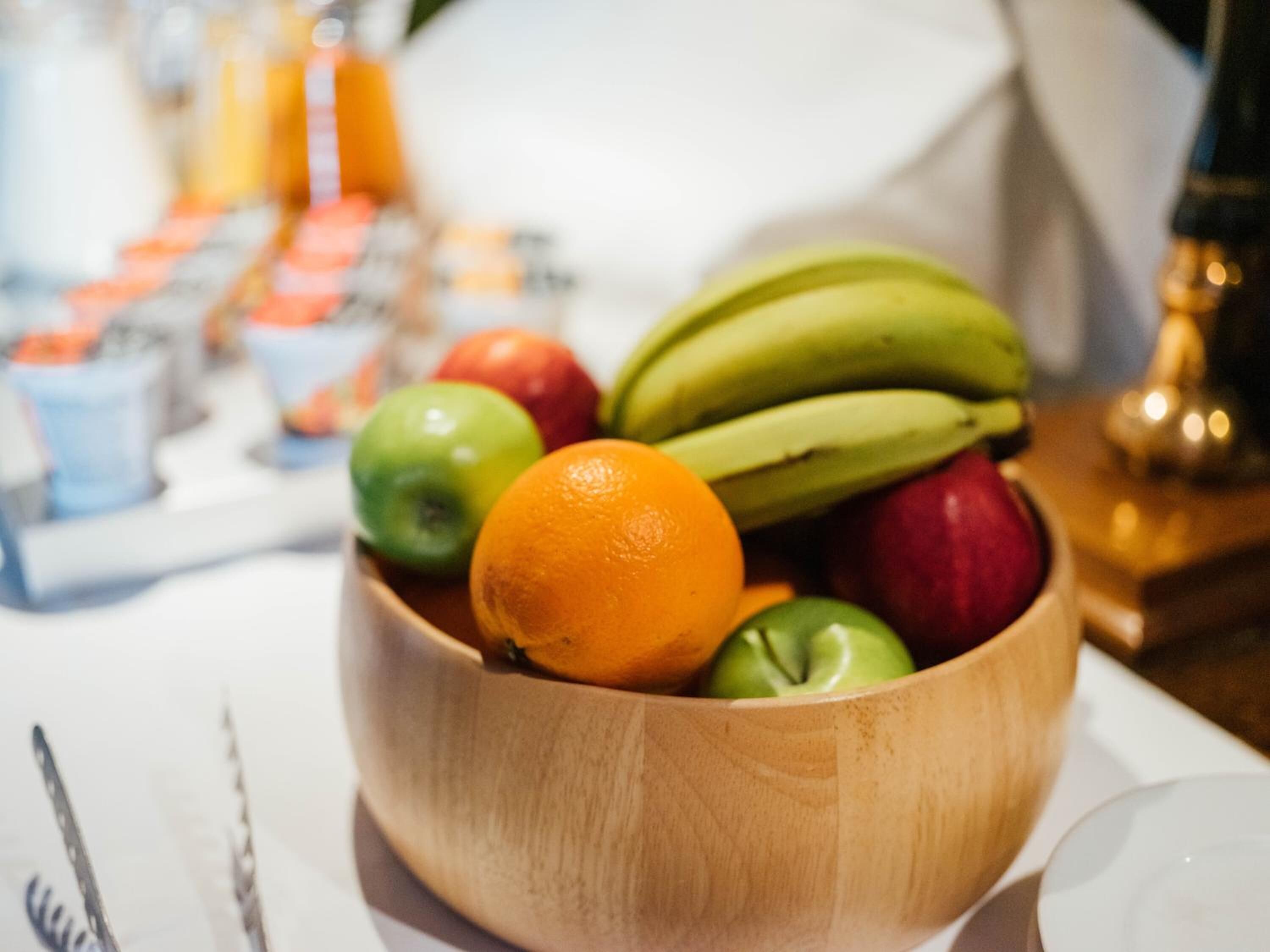 A wooden bowl filled with fruits including bananas, apples, and an orange, placed on a table with a white cloth.