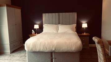 A neatly made bed with a grey headboard, two bedside tables with lamps, a grey upholstered chair, and a wardrobe.