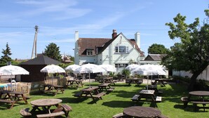 Een terras buiten met picknicktafels en parasols, een groot gebouw op de achtergrond en een gazon.