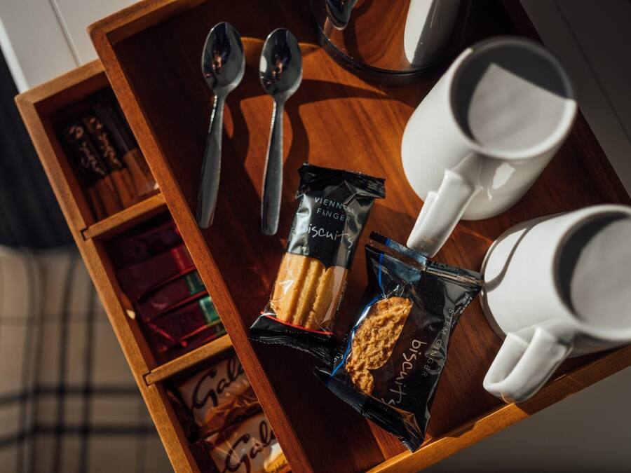 Una bandeja de madera con galletas, dos cucharas, dos tazas blancas y una pequeña caja de bombones.