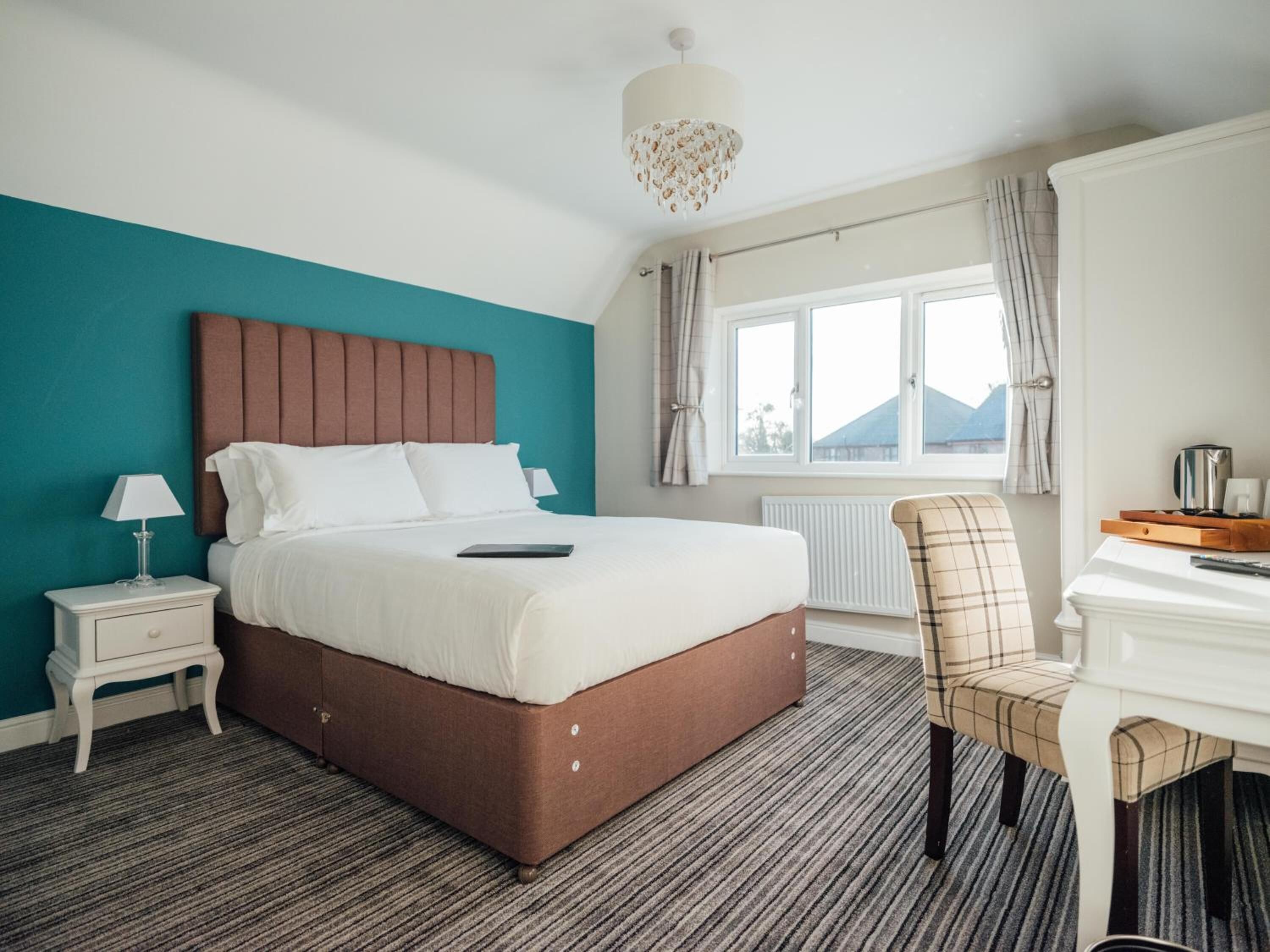A hotel room with a large bed, a bedside table with a lamp, a chair, a desk with a coffee maker, and a window with curtains.