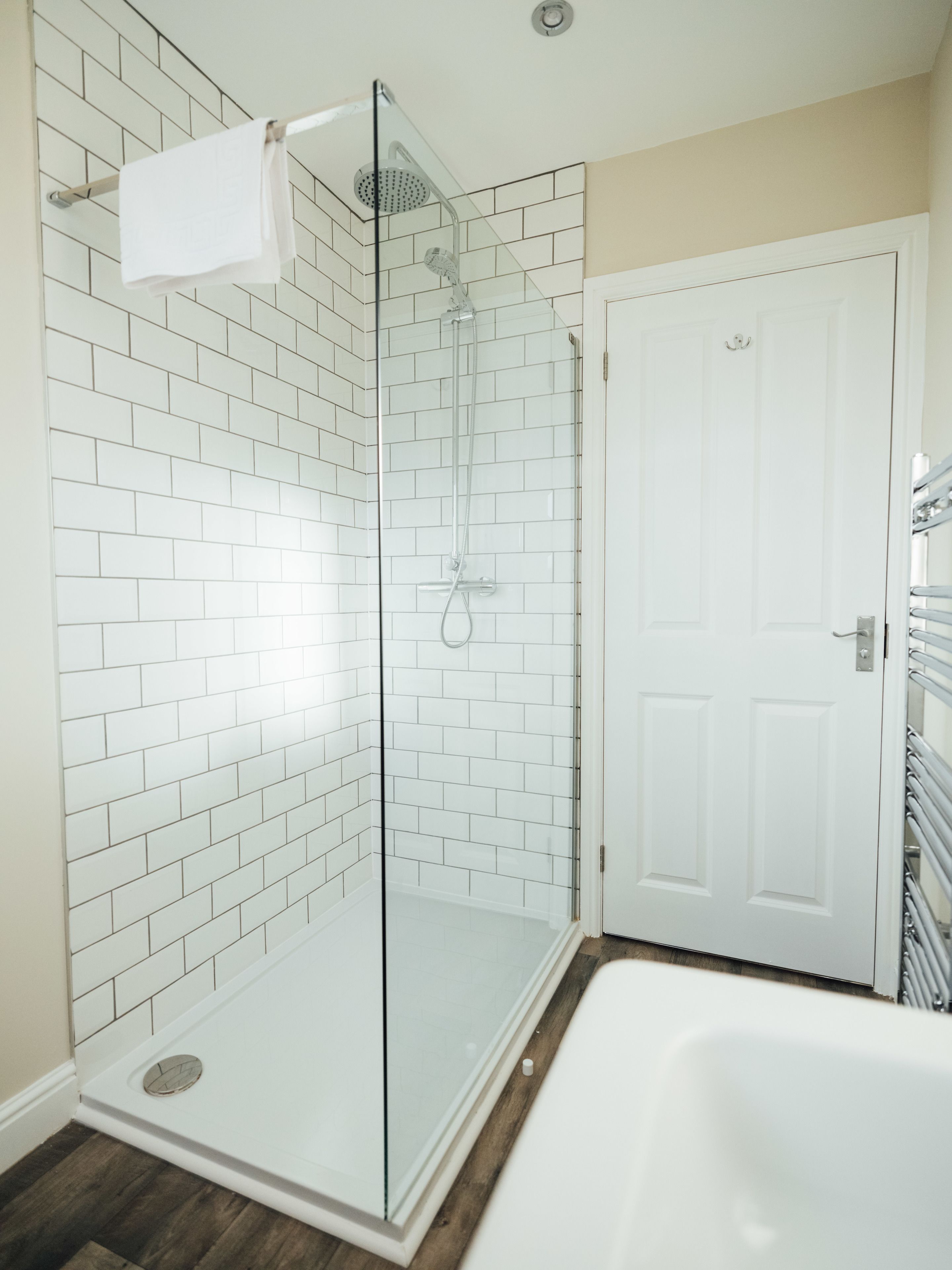 A modern bathroom with white subway tiles, a glass-enclosed shower, a white bathtub, and a wooden floor.