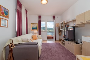 A compact hotel room with a sofa, a TV, a small kitchen area, and a balcony with a scenic view.