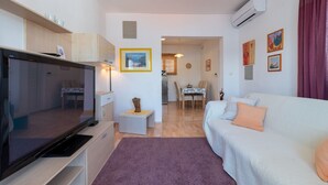 A modern living room with a flat-screen TV, a sofa, a dining table, and a kitchen area. - Mandic Apartment - Seaside Views, Beach Nearby (Mastrinka)