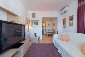 A modern living room with a flat-screen TV, a sofa, a dining table, and a kitchen area.