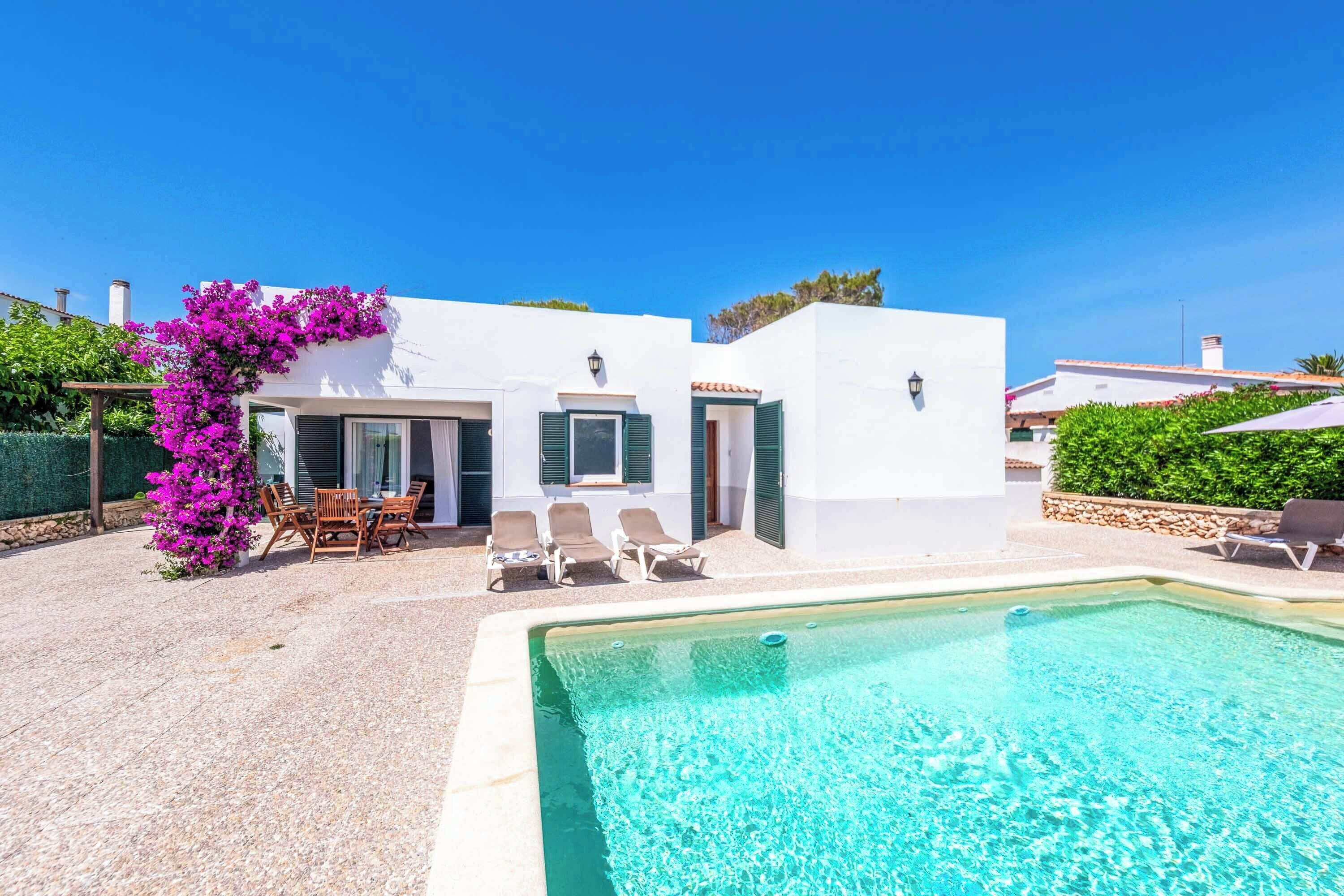 Una villa moderna con piscina, mobiliario exterior y una pérgola adornada con flores moradas.